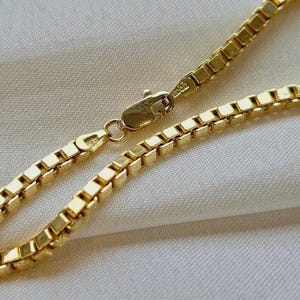 May include: Close-up of a gold-colored box chain necklace. The chain is made of small, square links and has a lobster clasp closure. The necklace is displayed on a white, textured surface.