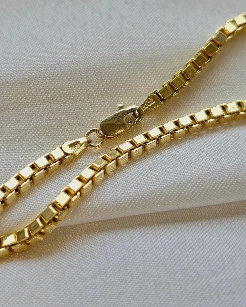 May include: Close-up of a gold-colored box chain necklace. The chain is made of small, square links and has a lobster clasp closure. The necklace is displayed on a white, textured surface.