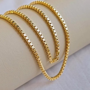 May include: A gold-colored box chain necklace. The chain is made of small, square links that create a smooth, continuous surface. The necklace is draped over a neutral-colored surface, highlighting its shine and design.