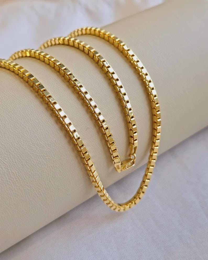 May include: A gold-colored box chain necklace. The chain is made of small, square links that create a smooth, continuous surface. The necklace is draped over a neutral-colored surface, highlighting its shine and design.