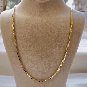 May include: A gold-colored necklace with a square link design is displayed on a mannequin. The necklace is draped around the mannequin's neck, showcasing its length and style. The background is neutral, highlighting the necklace's color and design.