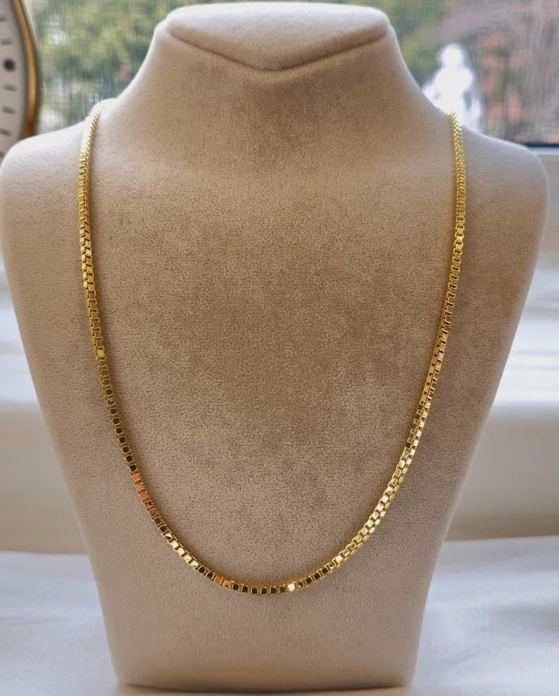 May include: A gold-colored necklace with a square link design is displayed on a mannequin. The necklace is draped around the mannequin's neck, showcasing its length and style. The background is neutral, highlighting the necklace's color and design.