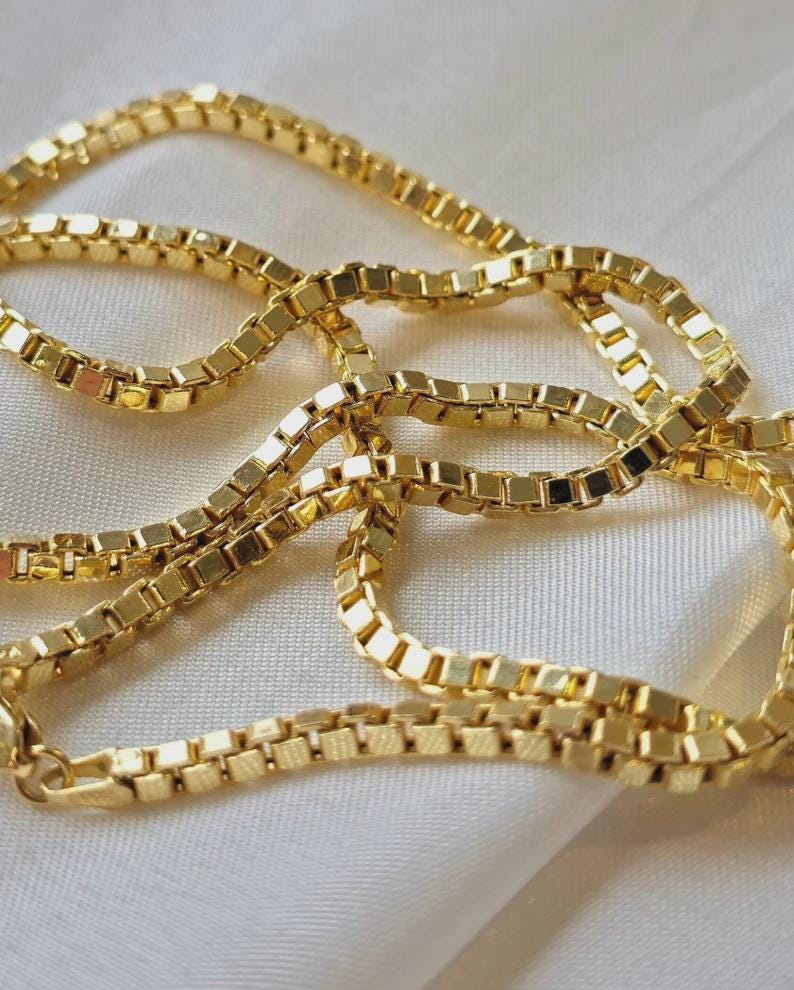 May include: A gold-colored box chain necklace. The chain is made of small, square links that are connected together. The necklace is lying on a white, silky surface. The chain is a bright, shiny gold color.