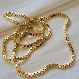 May include: A gold-colored box chain necklace. The chain is made of small, square links that are connected together. The necklace is lying on a white surface. The chain is a bright, shiny gold color.