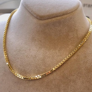 May include: A gold-colored box chain necklace displayed on a mannequin. The chain is made of small, rectangular links. The necklace is a bright, shiny gold color and is displayed against a neutral background.
