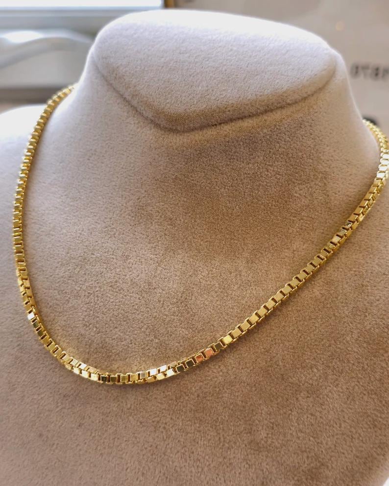 May include: A gold-colored box chain necklace displayed on a mannequin. The chain is made of small, rectangular links. The necklace is a bright, shiny gold color and is displayed against a neutral background.