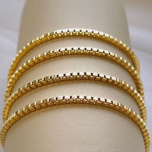 May include: Close-up of four gold-colored box chain necklaces. The chains are arranged in a curved pattern, showcasing their delicate, square-link design. The necklaces are displayed against a neutral, cream-colored background, highlighting their bright, reflective surface.