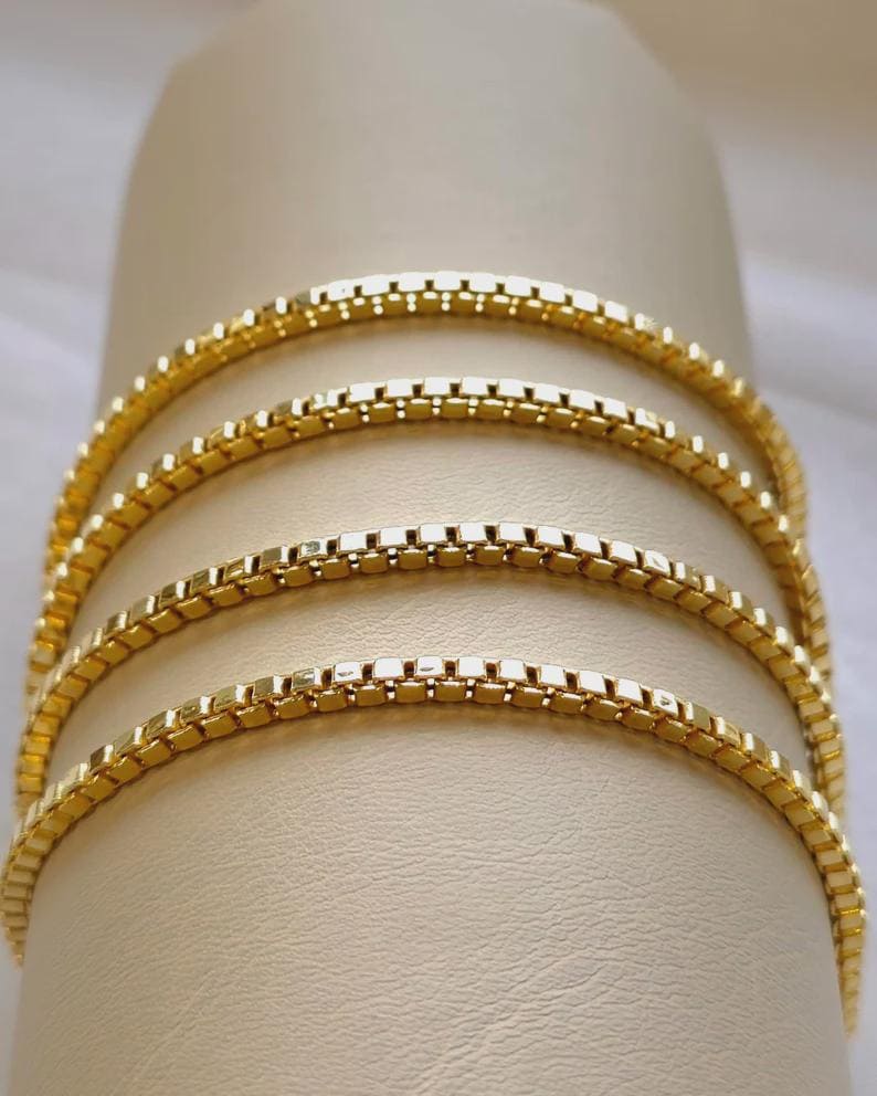May include: Close-up of four gold-colored box chain necklaces. The chains are arranged in a curved pattern, showcasing their delicate, square-link design. The necklaces are displayed against a neutral, cream-colored background, highlighting their bright, reflective surface.