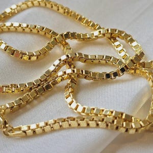 May include: A gold-colored box chain necklace. The chain is made of small, square links that are connected together. The necklace is lying on a white, silky surface. The chain is a bright, shiny gold color.