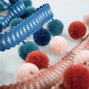 May include: Close-up of three different trims with pom-poms. The trims are blue, pink, and red. The pom-poms are made of soft, fluffy yarn.