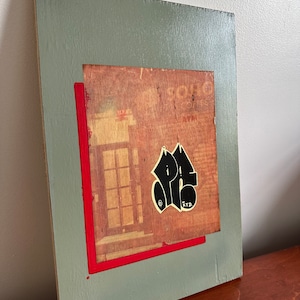 May include: A gray wooden frame with a red border around a vintage advertisement for "Solo" coffee. A black and white sticker with the word "PAIS" is on the advertisement.