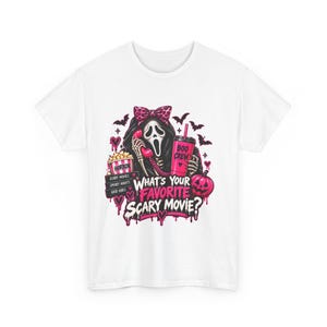 Puede incluir: Una camiseta blanca con un gráfico de una figura enmascarada con un lazo rosa de estampado de leopardo, sosteniendo un teléfono rosa y un vaso con la etiqueta BOO CREW. El diseño incluye palomitas, murciélagos, una calabaza y el texto WHAT'S YOUR FAVORITE SCARY MOVIE?