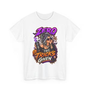 Puede incluir: Una camiseta blanca con un gráfico de una persona con el pelo revuelto y gafas de sol sosteniendo un vaso con un diseño de mano de esqueleto. El texto dice ZERO TRICKS GIVEN. La ilustración incluye una calabaza, murciélagos y telarañas en morado y naranja.