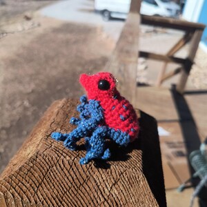 May include: A small, handmade crochet frog with a red body and blue legs. The frog has black eyes and is resting on a wooden surface. A unique, handcrafted item.