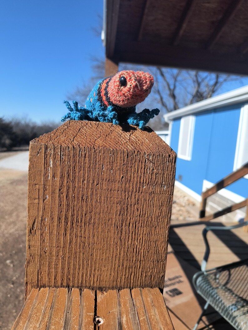 May include: A handmade crochet lizard in shades of blue, orange, and black rests on a wooden post. The lizard has a textured appearance with detailed stitching, and is positioned outdoors against a bright blue sky.