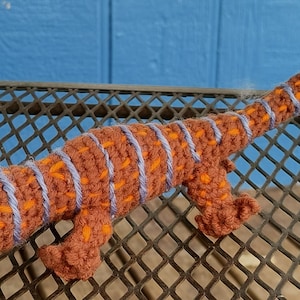 May include: A crocheted brown and orange lizard with blue stripes. The lizard has a yellow eye and is posed on a black metal grate.