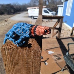 May include: A handmade crochet lizard in blue, orange, and black sits on a wooden post. The lizard has a blue body with orange spots, an orange head, and black stripes. The background includes a blue building and a sunny sky.