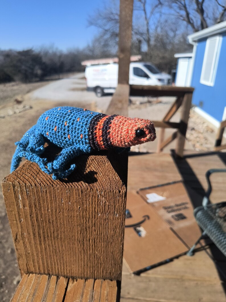 May include: A handmade crochet lizard in blue, orange, and black sits on a wooden post. The lizard has a blue body with orange spots, an orange head, and black stripes. The background includes a blue building and a sunny sky.