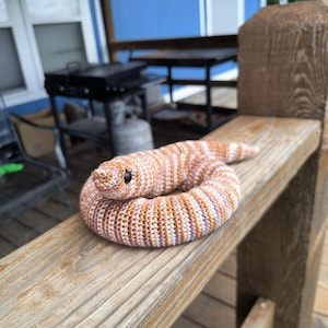May include: A coiled crochet snake toy featuring a striped pattern in shades of tan, white, and soft purple. The plush snake has a rounded head with small black bead eyes and a textured yarn surface, crafted with a consistent stitch throughout.