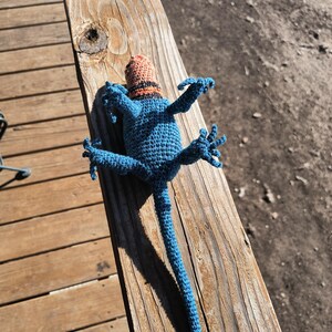 May include: A handmade blue crochet lizard toy with a small orange and black hat. The lizard is lying on a wooden surface, with its arms and legs outstretched. The toy is detailed and whimsical.