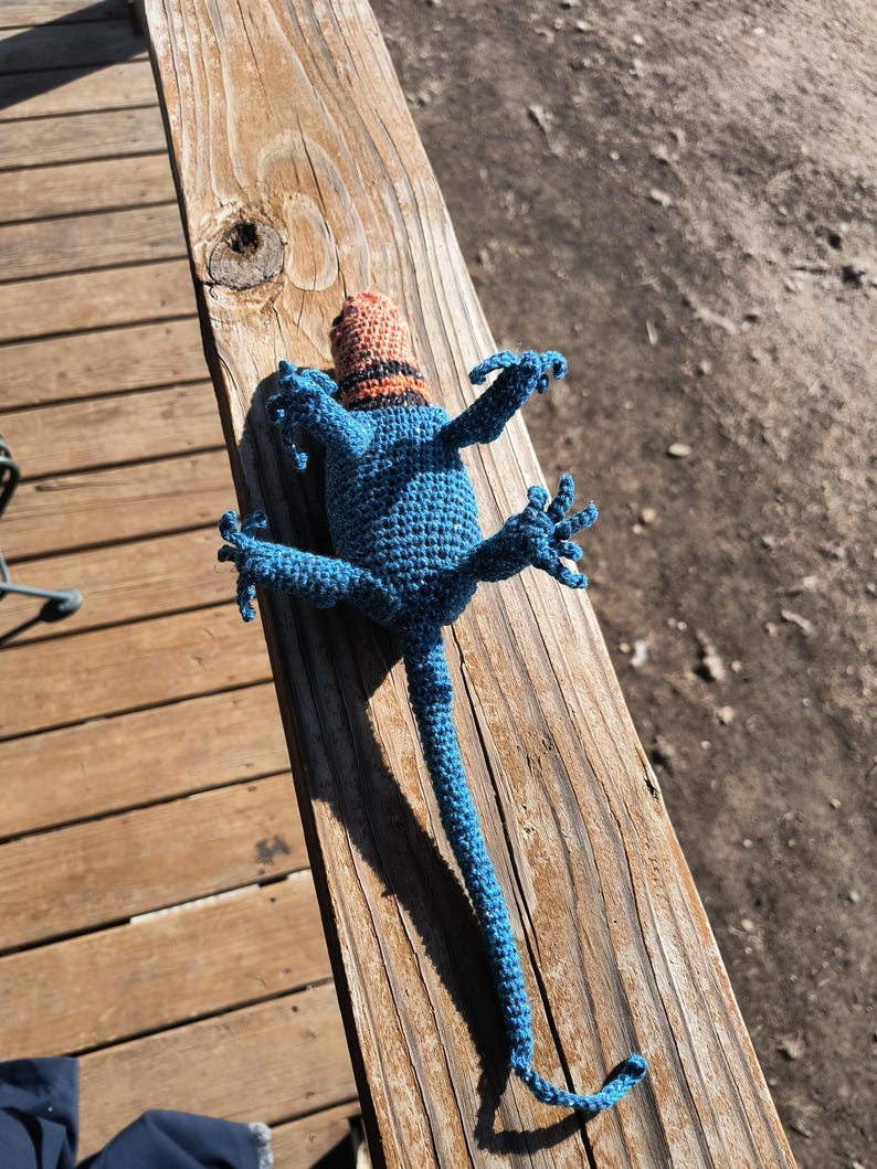 May include: A handmade blue crochet lizard toy with a small orange and black hat. The lizard is lying on a wooden surface, with its arms and legs outstretched. The toy is detailed and whimsical.