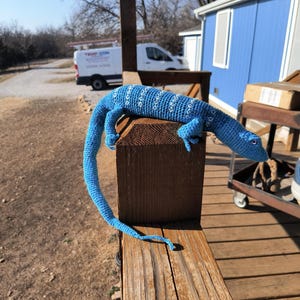 May include: A handmade blue crochet lizard with white patterned accents, resting on a wooden post. The lizard's long tail extends down, showcasing the detailed craftsmanship of this unique decorative item.