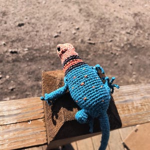 May include: A handmade crochet lizard in shades of blue, orange, and black. The lizard has a detailed pattern with orange spots on its back and a striped head. It is posed on a wooden surface.