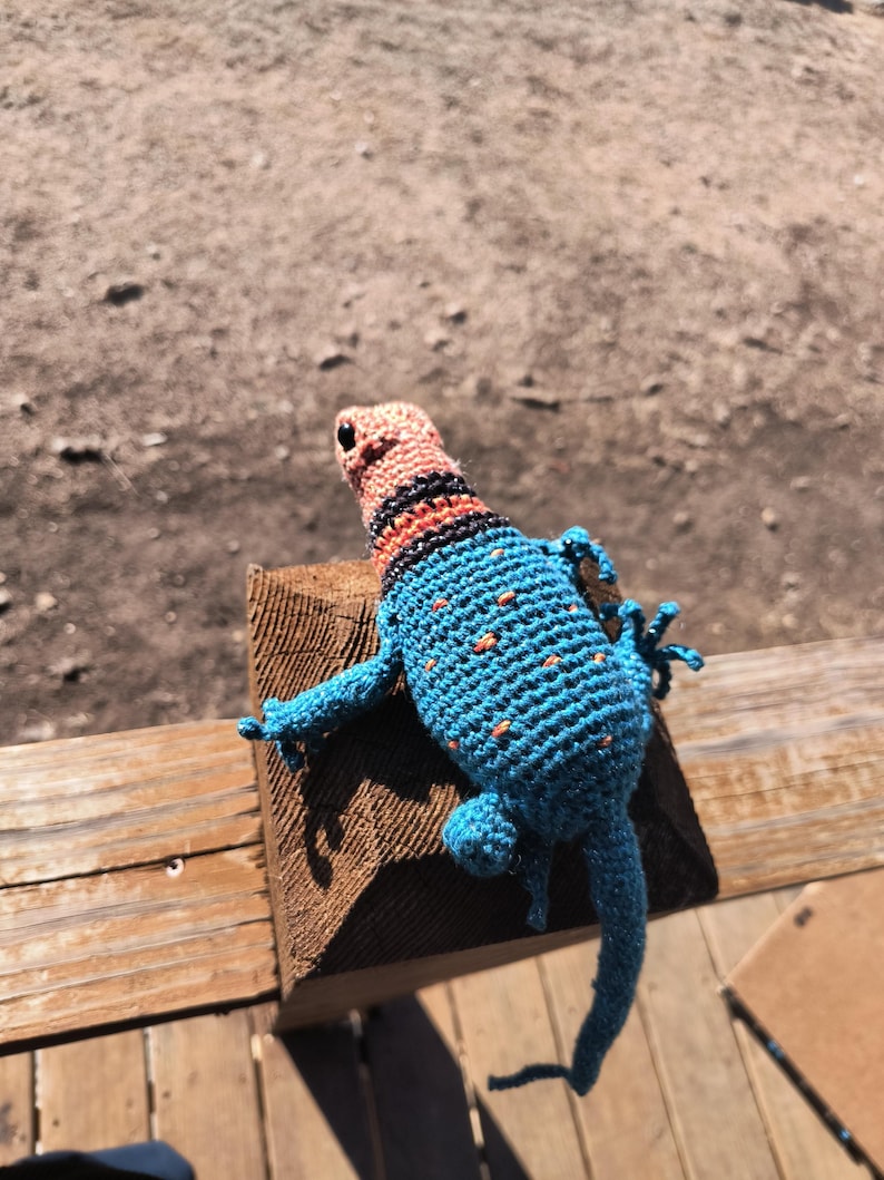May include: A handmade crochet lizard in shades of blue, orange, and black. The lizard has a detailed pattern with orange spots on its back and a striped head. It is posed on a wooden surface.