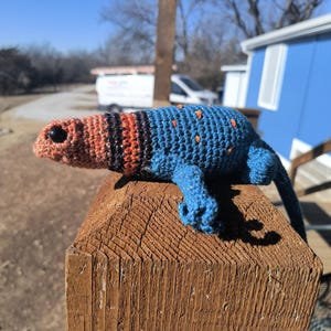 May include: A handmade crochet lizard in shades of blue, orange, and black, with small orange dots. The lizard is perched on a wooden post, showcasing its detailed craftsmanship and vibrant colors. The lizard's head is orange, transitioning to black and orange stripes.