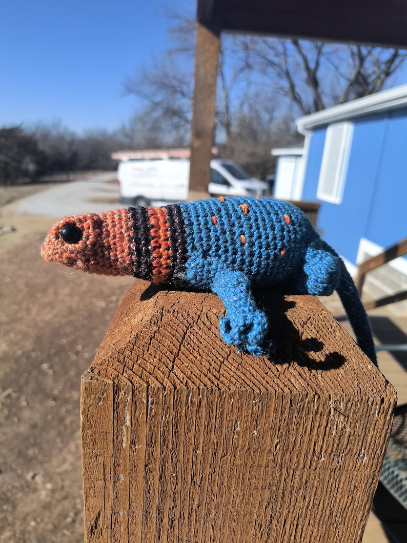 May include: A handmade crochet lizard in shades of blue, orange, and black, with small orange dots. The lizard is perched on a wooden post, showcasing its detailed craftsmanship and vibrant colors. The lizard's head is orange, transitioning to black and orange stripes.