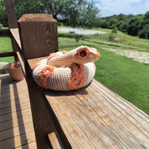 May include: A coiled crochet snake toy featuring a cream-colored body with vibrant orange patterned patches along its back. The snake has a textured, stitched surface and prominent, dark red, glossy eyes that stand out against its light head.