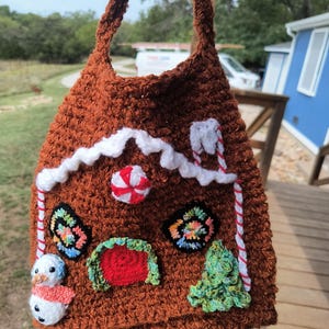 May include: A brown crocheted gingerbread house-shaped tote bag with white icing accents, a red door, and colorful details. The bag features a snowman, candy cane stripes, and a peppermint candy. The bag has a handle for carrying.
