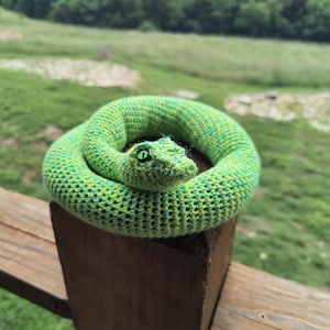 May include: A coiled, crocheted snake in shades of green and yellow rests on a wooden post. The snake's head is visible, featuring green eyes. The background shows a blurred green landscape.