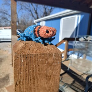 May include: A handmade crochet creature with a blue body, orange head, and black stripes. The amigurumi is perched on a wooden post, showcasing its detailed craftsmanship and vibrant colors. The creature has small, detailed legs and a textured appearance.