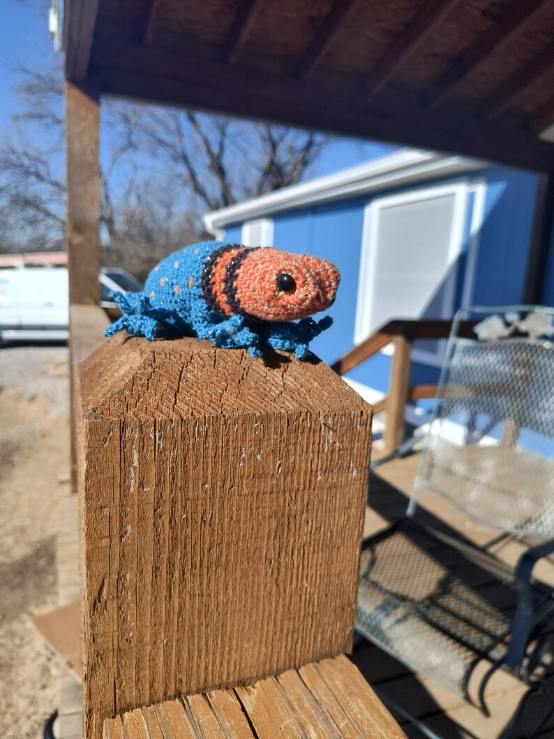May include: A handmade crochet creature with a blue body, orange head, and black stripes. The amigurumi is perched on a wooden post, showcasing its detailed craftsmanship and vibrant colors. The creature has small, detailed legs and a textured appearance.