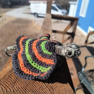 May include: A handmade crochet turtle with a shell featuring orange, green, and black stripes. The turtle's head and body are a mix of gray and brown yarn. The shell has silver glitter accents. The turtle is sitting on a wooden surface.