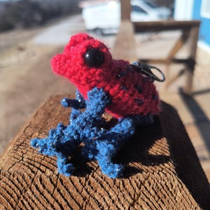 May include: A handmade crochet frog figurine with a red head and blue body. The frog has large black eyes and is sitting on a wooden surface. The frog is a keychain.