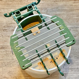 Op de afbeelding: Een groene en zilveren plantenpot houder met de tekst "DELLA PLANTS" en "PLANTA POTTY". De houder is geplaatst in een witte emmer. De houder heeft een rechthoekige vorm met meerdere sleuven voor planten.