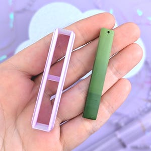 May include: A light purple rectangular open-frame cutter and a solid olive green rectangular bar with a small hole at the top. The cutter has a hollow center, while the green bar features a smooth, matte finish and a uniform, elongated shape.