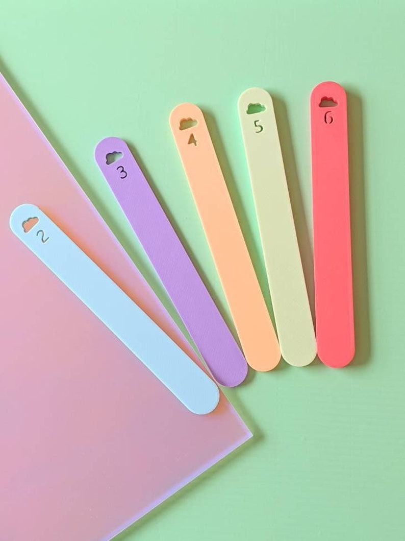 May include: Six colorful wooden sticks with cloud-shaped tops, each numbered 2 through 6. The sticks are arranged in a fan shape on a pink and green background.