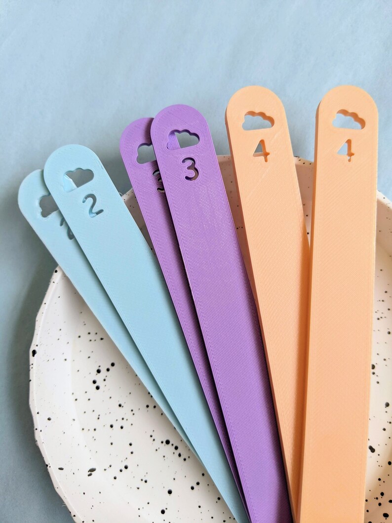 May include: Five colorful plastic bookmark strips with cloud cutouts and numbers 1 through 4. The bookmarks are arranged in a fan shape on a white speckled surface.