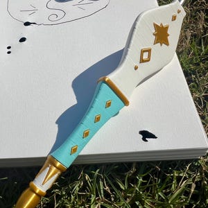 May include: A dip pen with a white upper handle featuring gold star and diamond accents, a turquoise lower handle with gold diamond inlays, and a gold pointed tip. The pen rests on a white paper sheet with a hand-drawn circle and ink spots.