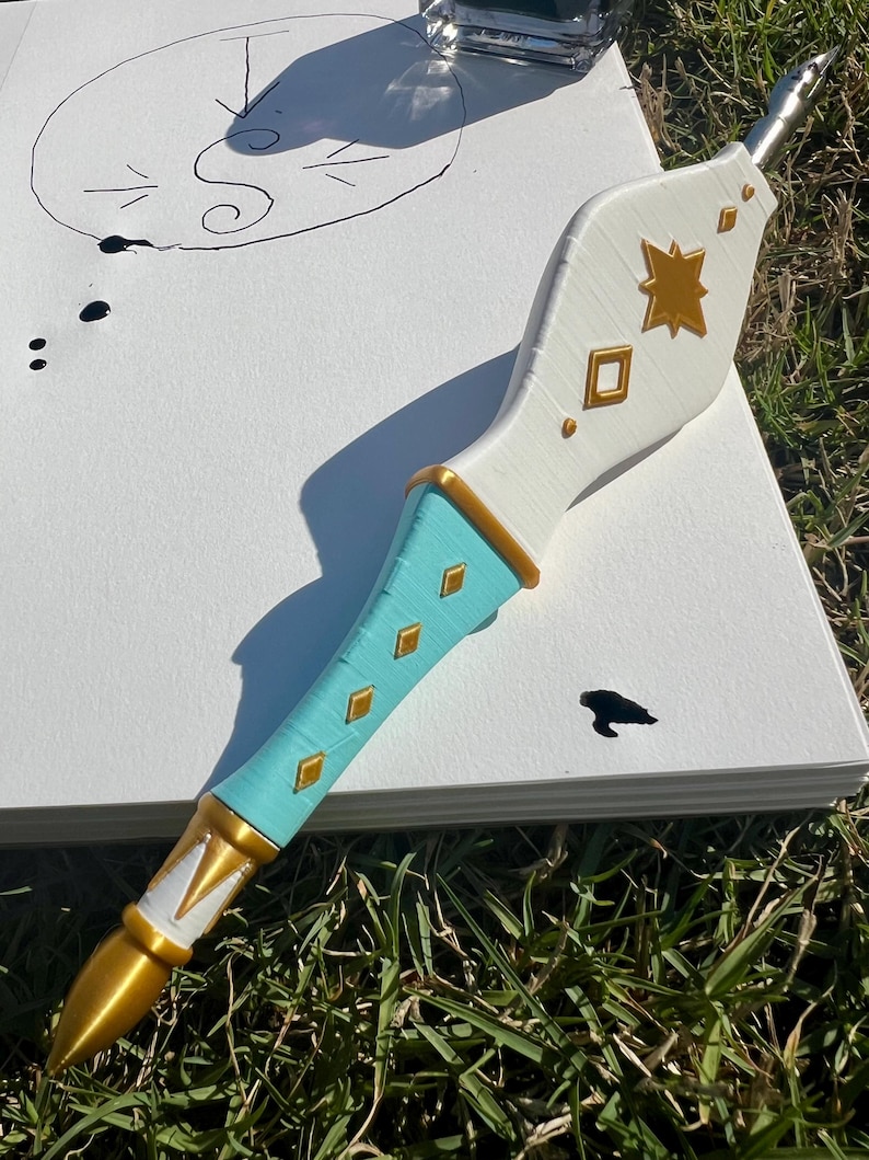May include: A dip pen with a white upper handle featuring gold star and diamond accents, a turquoise lower handle with gold diamond inlays, and a gold pointed tip. The pen rests on a white paper sheet with a hand-drawn circle and ink spots.