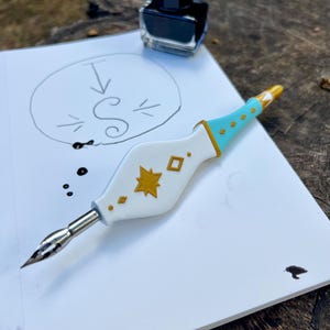 May include: A white and light blue calligraphy pen with gold star and diamond accents, featuring a metal nib. The pen rests on a white sheet of paper with a hand-drawn letter S inside a circle, accompanied by a small glass bottle of black ink.