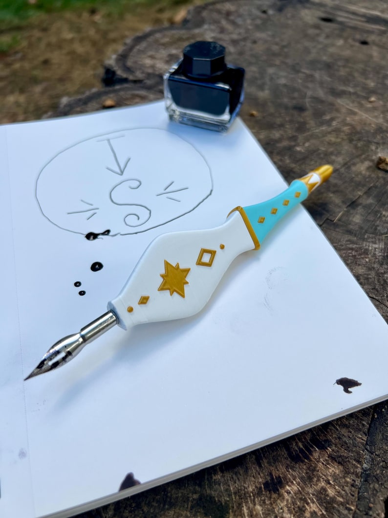 May include: A white and light blue calligraphy pen with gold star and diamond accents, featuring a metal nib. The pen rests on a white sheet of paper with a hand-drawn letter S inside a circle, accompanied by a small glass bottle of black ink.