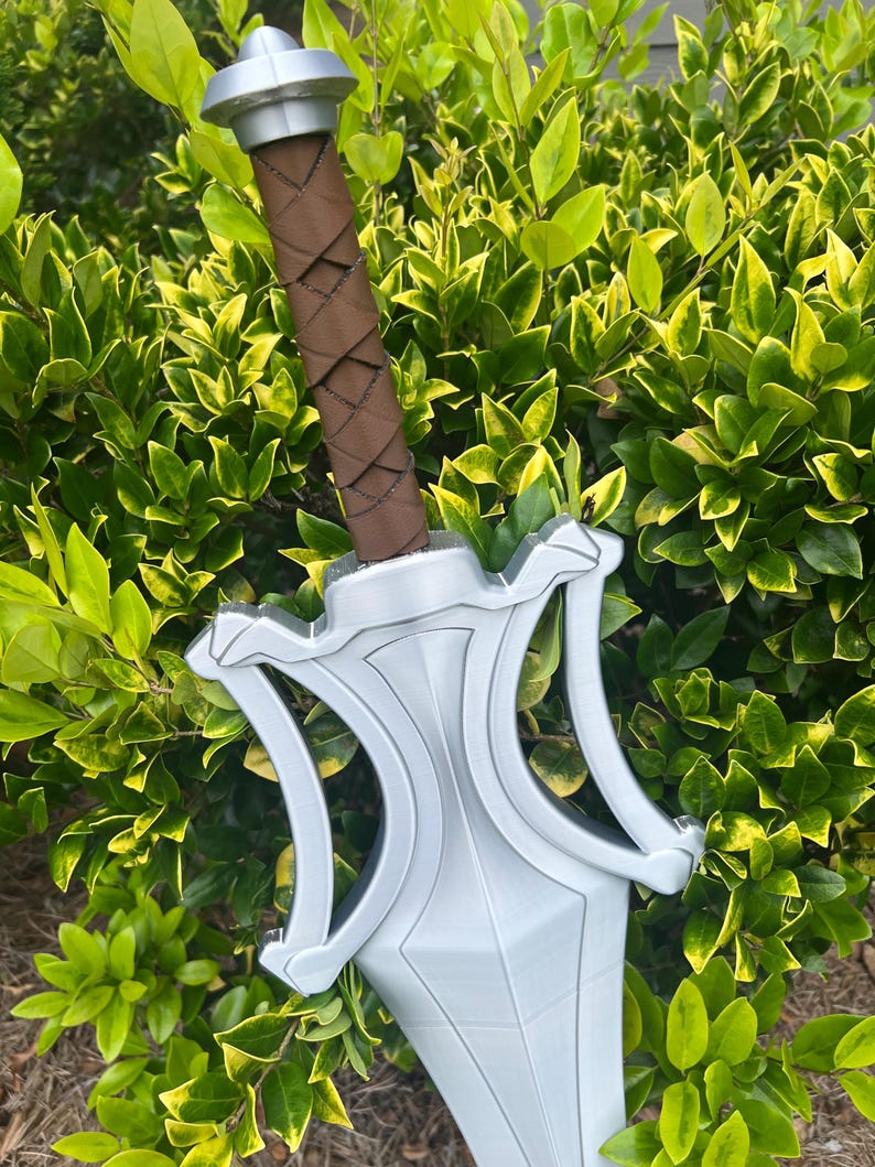Puede incluir: Una espada de color plateado con una guarda ancha y angular y un pomo. El mango est&aacute; envuelto en material marr&oacute;n con un patr&oacute;n entrecruzado. La hoja es ancha y se estrecha hacia la punta, mostrando un acabado met&aacute;lico liso en todo el cuerpo.