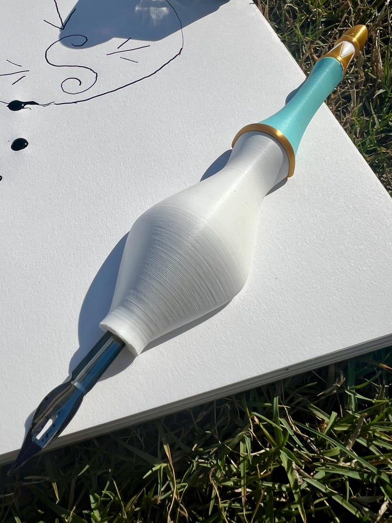 May include: A calligraphy pen featuring a white, bulbous, 3D-printed handle with a smooth texture. The upper section of the handle is light blue with a gold-colored ring accent. A silver metal nib is inserted into the base of the white handle.