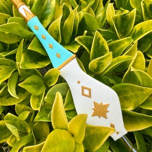 May include: A decorative wand featuring a pointed gold tip, a light blue handle with gold diamond-shaped accents, and a wide white base adorned with a gold star and diamond-shaped details. The object has a metallic nib at the bottom end.