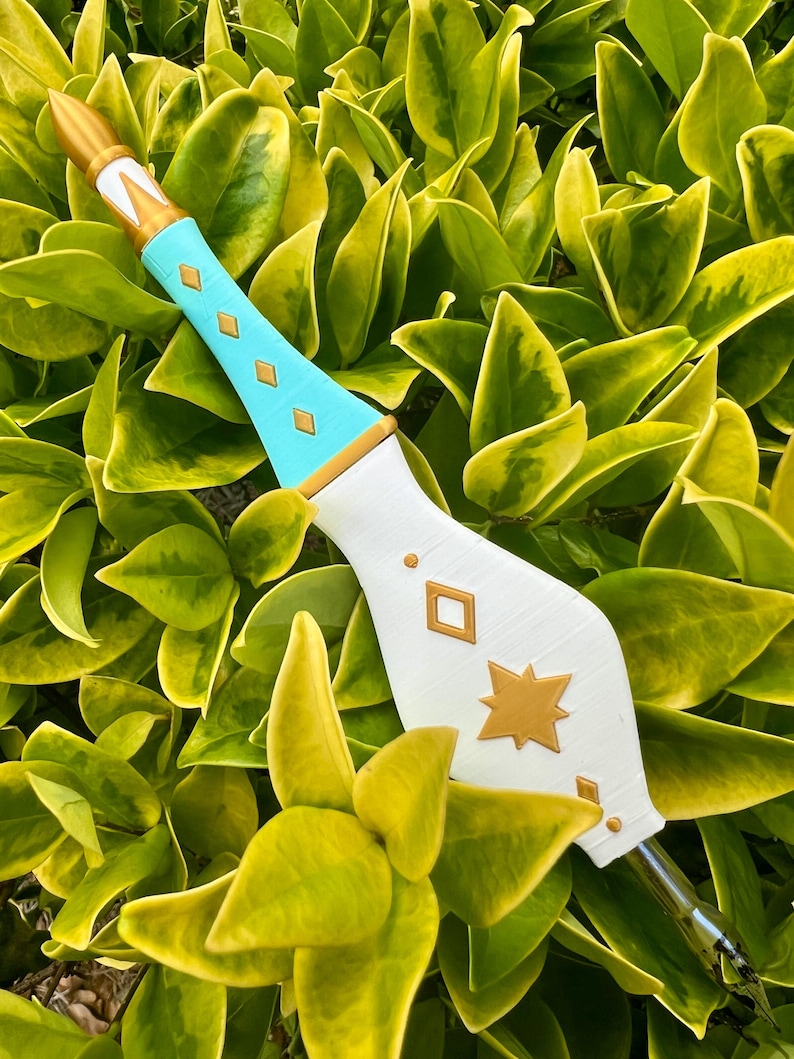 May include: A decorative wand featuring a pointed gold tip, a light blue handle with gold diamond-shaped accents, and a wide white base adorned with a gold star and diamond-shaped details. The object has a metallic nib at the bottom end.