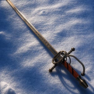 Puede incluir: Una espada ornamentada con una empuñadura de bronce y un mango marrón rojizo. La hoja plateada tiene grabados. La espada está sobre nieve, con un diseño de guarda detallado. Longitud estimada: 90 cm.